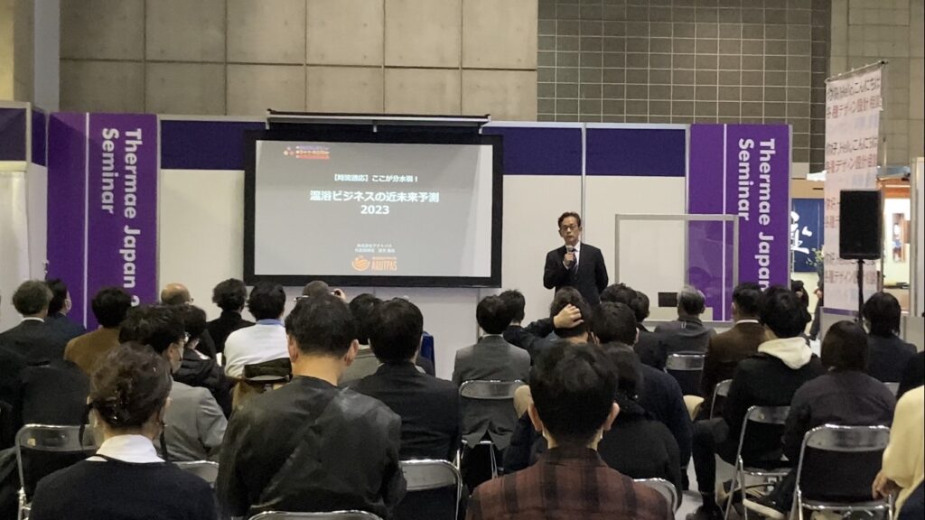 温浴ビジネスの近未来予測2023というスライドを背景にセミナーで講演する男性の様子。