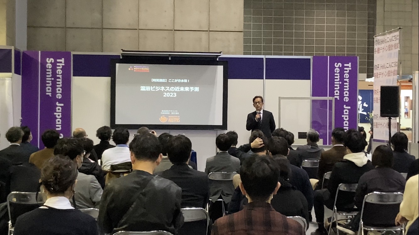 温浴ビジネスの近未来予測2023というスライドを背景にセミナーで講演する男性の様子。