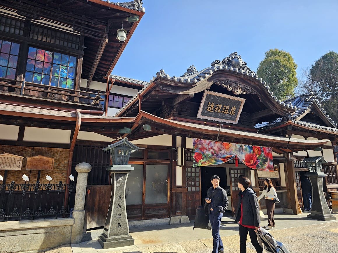 歴史ある木造建築の道後温泉本館の正面玄関前を歩く観光客の人々。