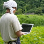 施設の屋外で頭にタオルを巻いた人物が、ノートパソコンを操作して業務を行っています。