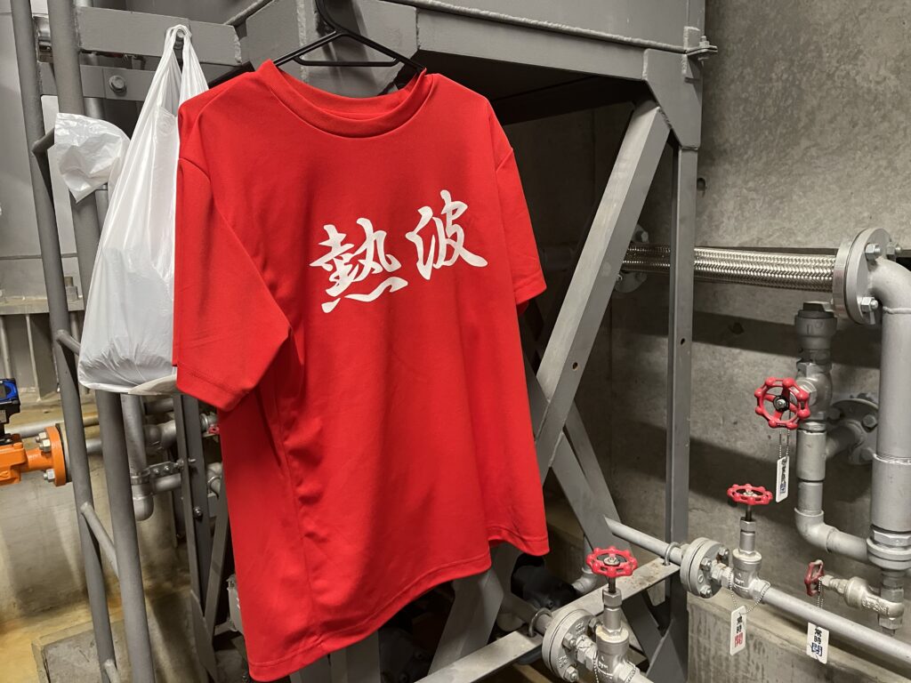温浴施設のバックヤードにある配管の前に、熱波と書かれた赤いTシャツがハンガーで掛けられています。