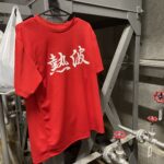 温浴施設のバックヤードにある配管の前に、熱波と書かれた赤いTシャツがハンガーで掛けられています。