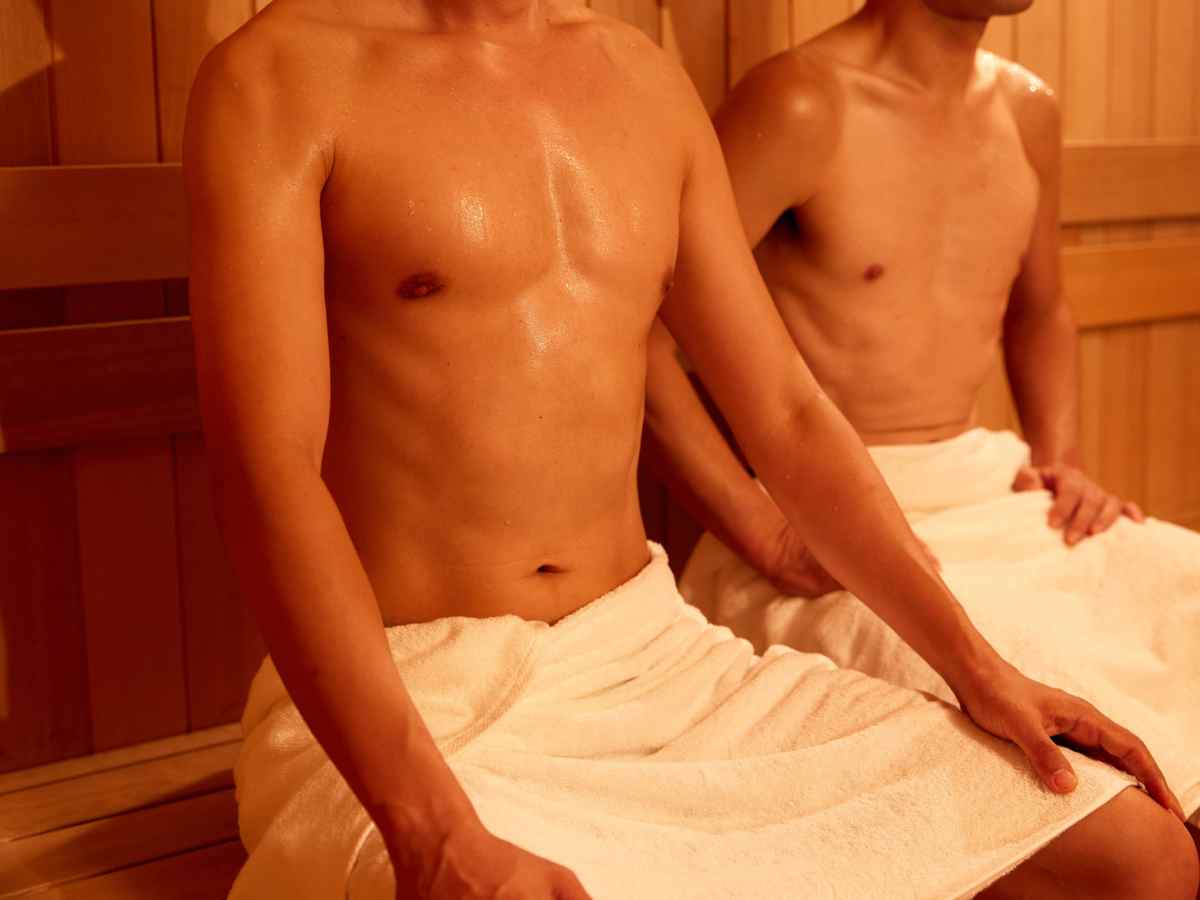 サウナ室内のベンチで腰に白いタオルを巻き、汗を流しながら並んで座る2人の男性の様子