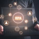ノートパソコンを持つ人物の手元とRPAやデータ処理を表すネットワークアイコンのグラフィックが表示されています