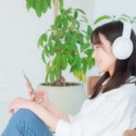 白いヘッドホンを装着した女性が、スマートフォンを持ちながら笑顔で座っています。