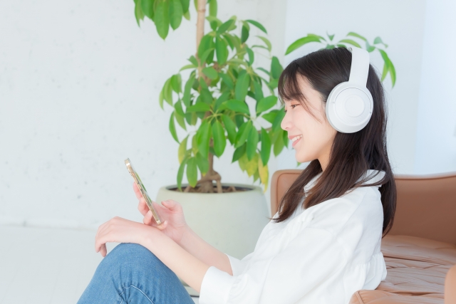 白いヘッドホンを装着した女性が、スマートフォンを持ちながら笑顔で座っています。