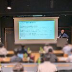 セミナー会場でスライドを背に登壇者がマイクを持って講義を行う様子。