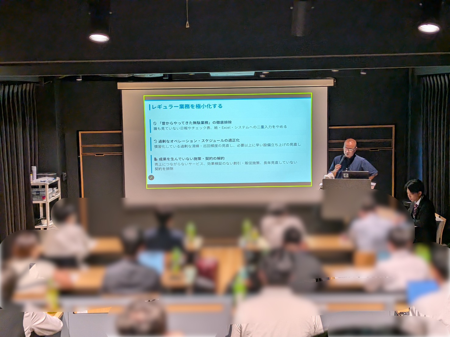 セミナー会場でスライドを背に登壇者がマイクを持って講義を行う様子。